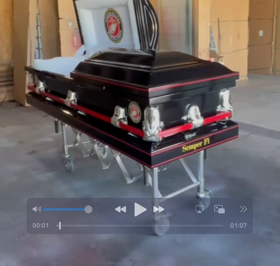 The United States Marine Corp USMC Casket in black with red trim and silver handles rests on a collapsible stand. Its partially open lid reveals a Marine Corps emblem and "Semper Fi," showing clear USMC pride. Video player controls are visible.