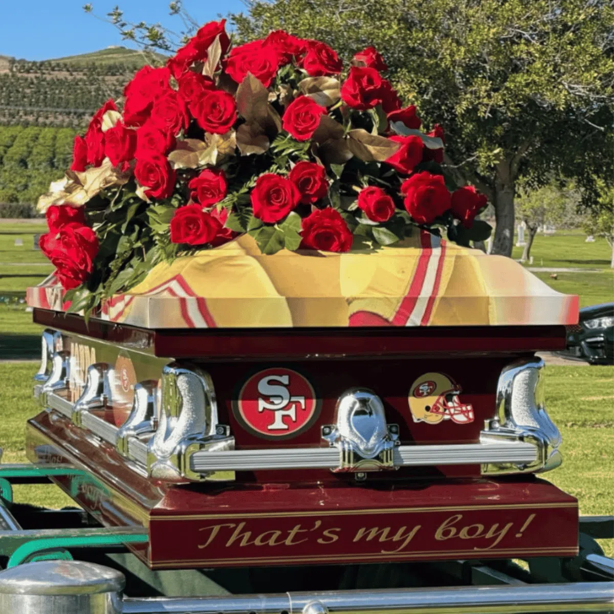 A maroon casket decorated with San Francisco 49ers logos and the phrase Thats my boy! is topped with a large bouquet of red roses in an outdoor cemetery setting.